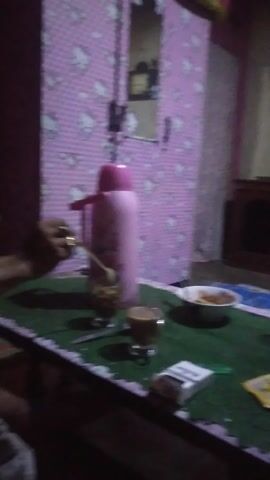 The video depicts an indoor scene, likely a home, in Kecamatan Cimaung, Indonesia. The lighting is dim, suggesting it is evening or night.

The initial frame (0:00-0:02) shows a room set up with two tables. The table on the right is covered with a green and blue patterned tablecloth and holds various items: a pink thermos, a clear glass with a light-colored beverage, a white bowl containing food, a small yellow packet, a cigarette pack, and two cups with a dark beverage, possibly coffee or tea. In the background, a wooden cabinet with a television set on top is visible. To the left, another table with a pink and white patterned tablecloth is partially visible.

From 0:02 to 0:04, a woman is seen sitting on the floor, smiling and looking towards the camera. She is wearing a red t-shirt and dark pants. The floor is covered with a multi-patterned rug. Behind her, a small white appliance, possibly a rice cooker, is plugged in. The scene appears casual and relaxed. There is no clearly discernible text in the video.