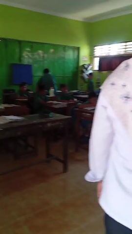 The video shows a classroom with students sitting at desks and a teacher standing. The classroom walls are painted green, and there is a window on the right side of the room. The students are wearing green uniforms, and they appear to be engaged in a lesson. The teacher is also wearing a green uniform and is interacting with the students. The overall atmosphere of the classroom appears to be calm and focused.