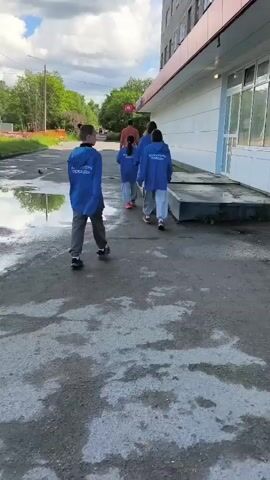 A group of children in blue jackets walks away from the camera on a paved path. The jackets have writing on the back in white text. The path is wet with puddles of water reflecting the sky. The children are walking next to a building with a white facade and a red trim above the windows. Trees and construction fencing are visible in the background under a cloudy sky. The camera is positioned at a low angle, following the children.