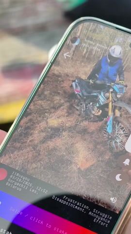 A hand holds up a smartphone displaying a video of a motorcyclist struggling in a muddy forest. The video appears to have been posted online, as information is shown in an overlay: a username "iloveyou," a date "03/28/2026," a timestamp "12:52," and a location "Goryachii Les, Belarus." Other text includes "Frustration, Struggle, Disappointment, Annoyance, Effort" and instructions to "swipe/click to stake." The environment visible in the phone screen shows a close-up view of the muddy ground and surrounding foliage. The focus remains on the phone screen and its contents.
