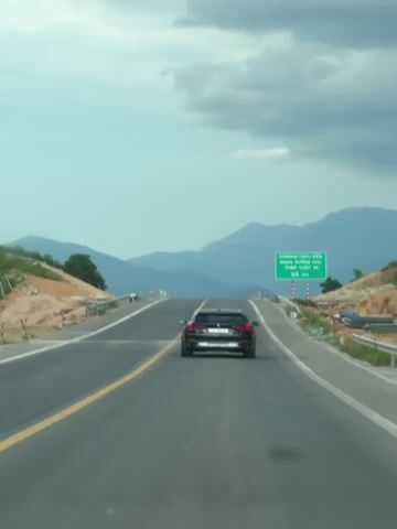This is a first-person perspective video of a black SUV driving on a highway. The car is traveling uphill and appears to be the only vehicle in close proximity. The highway has two lanes in each direction, separated by a yellow line. There are mountains in the background under a cloudy sky. A green road sign is visible on the right side of the road. There are some construction materials and debris on the side of the road. A person is walking on the left side of the highway in the distance.