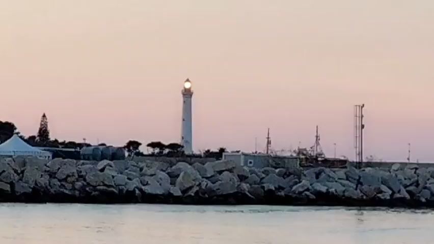 This media file captures a serene coastal scene at dusk, likely near San Vito Lo Capo, Italy. The primary focus is a tall, white lighthouse standing prominently on a rocky breakwater. The lighthouse, a classic cylindrical structure, has its lantern room illuminated, casting a faint glow against the fading light of the sky.

The breakwater itself is composed of large, irregular gray rocks, forming a sturdy barrier against the gentle waves of the sea that occupy the foreground. Behind the lighthouse and the breakwater, a collection of structures and natural elements are visible. To the left, a white tent structure, possibly a temporary canopy, suggests human activity or facilities at the location. Further back, trees, including what appears to be a cypress tree, dot the landscape, adding a touch of greenery. Other smaller buildings and structures, as well as some masts or poles, are scattered along the breakwater, hinting at a harbor or port area. The sky transitions from a pale pink to a soft purple, indicating the twilight hours, with no clouds visible, suggesting calm weather.

The overall mood of the image is peaceful and tranquil, evoking a sense of calm as the day draws to a close. The combination of the lighthouse, the sea, and the soft evening light creates a picturesque and slightly romantic atmosphere. There is no visible text in the media file.