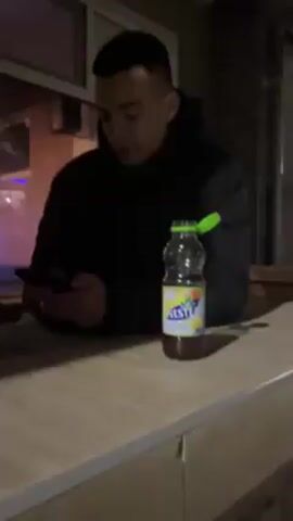 A person is standing indoors, looking at a phone. A bottle of Schweppes is on the counter. Two candle-like lights are visible through the window. The person is wearing a black and white jacket.