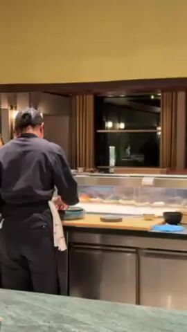 Here's a description of the video:

The video shows a sushi chef preparing food behind a sushi counter. The chef is wearing a dark uniform and a cap. He has a towel tucked into his apron. He appears to be arranging ingredients on a plate. Behind him is a glass display case with various sushi ingredients. The counter is made of light-colored wood. The restaurant has a modern, upscale design. A green marble countertop is visible in the foreground.