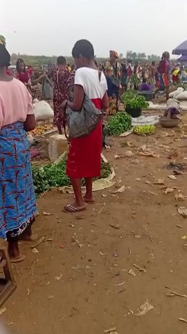 The video shows a busy marketplace. There are many people, mostly women, walking around and interacting with each other. Some are selling goods, including greens and bananas, laid out on the ground. The ground is dirt and is strewn with trash and debris. There are also several white bags, possibly containing cement, lying around. A blue umbrella provides shade for one of the vendors. The sky is overcast.