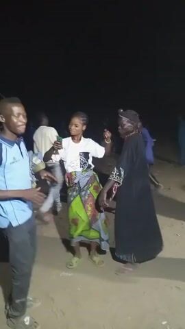 The video, taken at night, shows a group of people dancing in an outdoor setting that appears to be a dirt field or open area.  At least eight people are visible. A woman in a white t-shirt and brightly colored printed skirt holds a phone, possibly recording. She interacts with an older woman in a long black gown.  A man in a light blue polo shirt with dark sleeves dances nearby, holding what appears to be folded clothing. Other people are seen in the background, some wearing casual clothing. The environment is dimly lit.  The camera moves, following the action and providing varying views of the dancing participants.