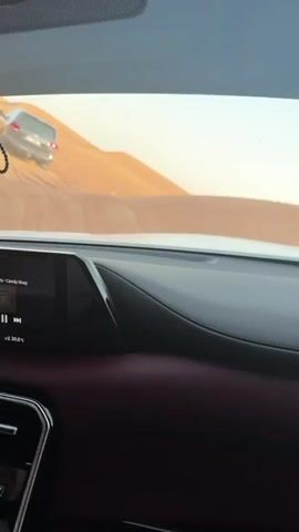 The video shows the dashboard of a vehicle, possibly a car, with a glimpse of red leather seats. Through the windshield, a large sandy desert landscape is visible under a bright sky. A silver SUV is driving up and over sand dunes. The car's movements are noticeable, and music plays in the background.