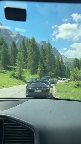 The video shows a view from inside a car driving on a road with a line of other cars ahead. The road is paved and bordered by green grassy slopes with numerous tall evergreen trees. In the background, large, rugged mountains rise under a bright blue sky with scattered white clouds. The lead car in the footage is a black convertible with its top down. The camera is positioned to capture the dashboard and windshield of the vehicle from which the video is recorded, revealing a GPS navigation screen on the lower left. The other vehicles are also mostly dark-colored SUVs and cars, suggesting a group outing or convoy. The scene appears to be a scenic mountain pass or road in a mountainous region during daylight.