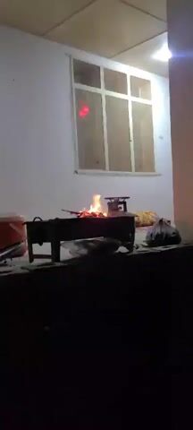 The video shows a nighttime scene of an outdoor cooking area. A small charcoal grill is visible with a fire burning inside. To the right of the grill, on the ground, are a plastic bag and some other items. In the background, there's a building with a window lit by an interior light. Two people are visible, crouching down and appearing to be preparing food on the ground near a blue container and bottles. The camera then pans out to show the dark surrounding outdoor area, suggesting a yard or open space. The ground appears to be sandy or light-colored.