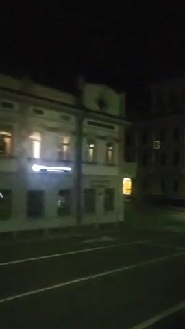 The video shows a nighttime street view with buildings on both sides. The street is paved and marked with white lines. Several streetlights are visible, illuminating the scene. The buildings appear to be old, with multiple floors and windows. Some windows are lit, suggesting occupancy. There is a crosswalk on the street. A car is visible in the lower right portion of the frame. The overall impression is that of an urban environment at night, with moderate lighting and no visible human activity.