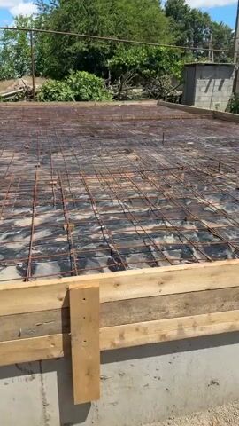 The video shows an outdoor construction site on a sunny day. The primary focus is on a concrete foundation being prepared. There is a wooden formwork in place, surrounding an area covered with black plastic sheeting and reinforced with a grid of metal rebar. The rebar is laid out in a regular pattern, indicating it's for structural support of the concrete slab. Beyond the foundation, the ground is dirt and sand. In the background, there are trees and vegetation, suggesting a rural or semi-rural setting. There are no people visible in the video, and no actions or interactions are occurring. The environment appears to be a work in progress, with construction materials and debris present.