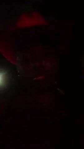 The video shows a person with dark skin and long braids, wearing a red cowboy hat and a necklace. The person is looking directly at the camera. The lighting is dim, but as the video progresses, the light brightens, revealing more of the person's features and clothing. The background is blurry and indistinct.