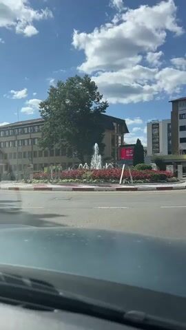 The video shows a circular intersection with a fountain in the center, surrounded by red flowers and green bushes. Several buildings are visible around the intersection, including a multi-story brown building with a dark sloped roof on the left, and taller, lighter-colored buildings on the right. One of the buildings on the right has the word "LUCA" in large yellow letters. A white SUV is driving around the intersection, and a silver car is parked on the right side of the road. The sky is blue with fluffy white clouds. The camera appears to be inside a vehicle, looking out through the windshield. The overall impression is a sunny day in a town or city.