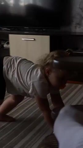 The video shows a young child with blonde hair in a white t-shirt and black leggings on all fours on a striped carpet. The child is facing away from the camera and appears to be reaching forward. In the background, there is a light-colored cabinet with a metal handle, a dark television, and a wall with textured wallpaper. The lighting suggests an indoor environment.