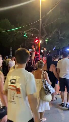 The video shows a nighttime outdoor gathering with a crowd of people standing and moving around. A DJ setup is visible on a raised platform, with speakers and lighting equipment. Several individuals are interacting with the DJ or observing the performance. The environment appears to be a street or public space, with trees and lighting overhead. The people are dressed in casual summer clothing, and the overall atmosphere suggests a lively event like a concert or festival.