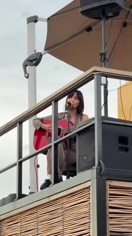 A young woman is performing live on a raised platform. She is sitting down, playing a red acoustic guitar, and singing into a microphone. She has short, dark hair and is wearing a tan suit. She is seated in front of a black speaker. The platform has a railing, and its side is covered in woven bamboo-like material. Above her is a canopy, supported by a white pole with a mechanical arm. The sky is overcast.