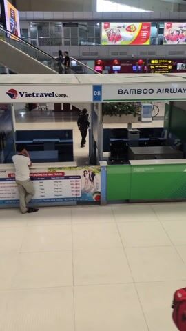 The video depicts the interior of an airport, likely in Vietnam based on the signage. The camera pans upward from a Vietravel Corp. kiosk, numbered 08, towards an "Information" booth numbered 07. A man is standing by the Vietravel kiosk, appearing to be on a phone call. Escalators can be seen in the background with people using them. Overhead signage reads "DA NANG INTERNATIONAL AIRPORT" in large, blue letters. Large digital billboards display advertisements. The floor is tiled, and the area appears well-lit. The overall impression is that of a bustling, modern airport.