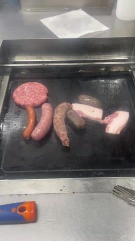 The video shows a flat top griddle with several pieces of raw meat cooking.  There's a hamburger patty, three different types of sausage, two pieces of bacon and two pieces of blood pudding. The griddle is hot and greasy, as evidenced by the steam rising off the food.  A spatula and a blue-and-orange object, possibly a tool, are on the stainless steel counter surface in front of the griddle. A white plate with some food scraps is also on the counter to the right of the griddle. Above the griddle, one can see a white sheet of paper and a roll of paper towels. Another griddle and a hamburger patty are faintly visible in the background.