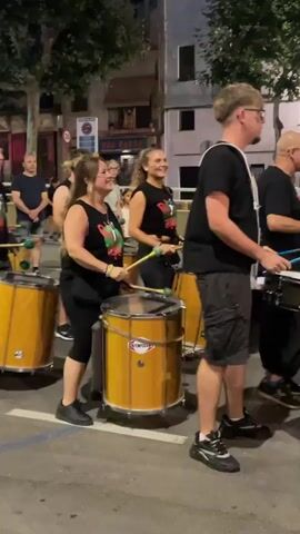 A group of people in black shirts are playing drums and percussion instruments in a street at night. The person in the foreground on the right is a young male with blond hair wearing glasses and a black t-shirt with a white strap across his chest and grey cargo shorts. He is holding drumsticks and looking to the right. To his left, an older bald man is playing a snare drum. Further to the left, a woman with brown hair is playing a large yellow drum. Another woman with blonde hair is also playing a drum behind her. In the background, there are buildings and trees. The street is paved and there are white lines on the ground.