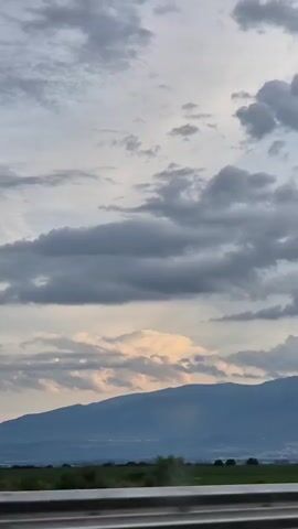 A car drives by on a highway. The sky is cloudy, with a mountain range in the distance. There is a green field in the foreground.