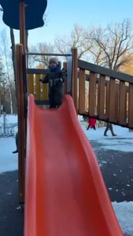 A small child wearing a warm winter outfit and hat is at the top of an orange playground slide. The child is holding onto the sides of the slide and looking down. Snow is visible on the ground around the playground equipment, and other people are walking in the background. The sky is clear and blue.