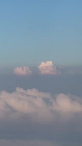 The video is a vertical shot of clouds from above. The sky is a pale blue, and the clouds are white and pink, illuminated by the sun. The clouds appear to be layered, with some closer and brighter than others. There are no people or objects visible in the video, only the clouds and the sky.