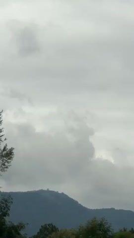 The video shows a highway scene with a view of lush green hills and trees on the right side of the road. The sky is overcast and appears to be cloudy. Several vehicles are visible on the highway, including a white truck with a red logo and a silver car. Orange markers are placed along the edge of the road. A speed limit sign indicating 80 is also visible. The camera is positioned inside a moving vehicle, likely a car, and the perspective shifts slightly as the vehicle moves. The overall impression is one of traveling through a scenic, rural landscape under a gray sky.