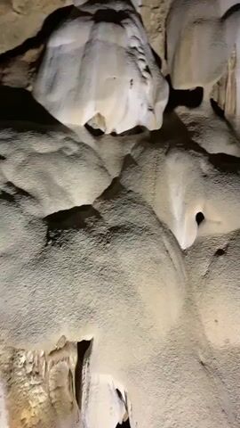 The video shows the interior of a cave. The camera pans across the ceiling and walls which are made of light-colored rock formations with crevices and rounded shapes.  There are a few people walking within the cave. One is holding up a phone, possibly recording. The lighting is dim, with artificial lights strategically placed to illuminate the rock formations.
