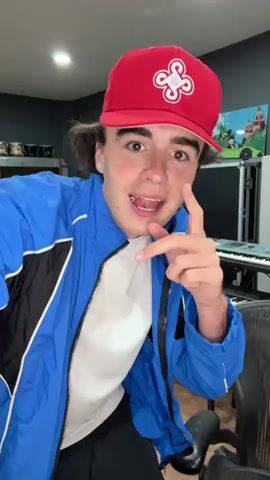A young male with dark hair, wearing a red baseball cap with a white logo, a blue and black jacket over a white t-shirt, and black pants with blue stripes, dances energetically in what appears to be a music studio. In the background, several keyboards are visible, along with a drum set and other audio equipment. The male gestures towards the camera and performs various dance moves, including pointing, making hand gestures, and swaying his body. The lighting is bright, illuminating the scene clearly.