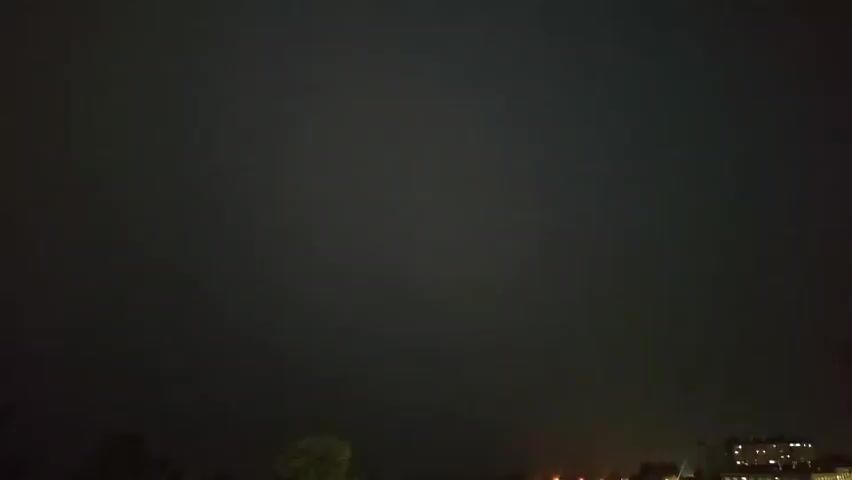 A brief flash of lightning illuminates a dark, cloudy sky over a city at night. The lightning strikes to the right of the frame, briefly outlining buildings and trees.