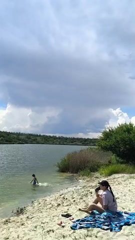 A wide shot shows a sandy beach next to a lake. In the foreground, a woman sits on a blanket, facing away from the camera, with her back to the lake. She is wearing a dark t-shirt and dark pants, and a black cap. To her left, on the sand, are a pair of flip-flops and what appears to be a second pair of flip-flops. Further back on the beach, near the edge of the trees, a man in blue shorts stands with his back to the camera, and a white car is parked. The lake has some reeds along the edge and appears to have some splashing near the left side of the frame. The sky is partly cloudy with areas of bright blue. The camera pans right, showing more of the beach, trees, and parked cars in the distance. A white SUV is visible under a tree. Further down the beach, a green chair sits on the sand. The pan continues right, showing more trees and a clear blue sky.