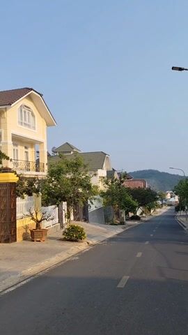 The video shows a straight asphalt road stretching uphill between houses under a clear, blue sky. A solitary mountain is visible in the distance at the center of the road's horizon. The houses lining the street are a mix of off-white and beige colors, most with green-tiled roofs and modest yards. Various trees and shrubs dot the yards and line the street. A black lamppost stands on the right, and an electrical pole with dangling wires is visible nearby. There are two people walking uphill together in the distance. The video is stationary.