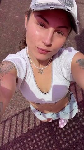 A woman with dark hair, fair skin, and visible tattoos is recording herself at a playground on a sunny day. She is wearing a white crop top, light-wash denim shorts, pink sneakers, and a distressed baseball cap. She also has a necklace with a decorative charm. The playground equipment is in the background and includes colorful metal structures and a red panel with various shapes. She looks at the camera and smiles slightly.