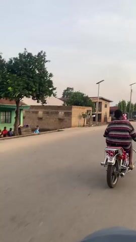 A first-person view from a moving vehicle shows a street scene. A person on a motorcycle is riding in front. On the left, a stone wall and a house are visible. On the right, a two-story building with laundry hanging from a line and a woman in a pink dress standing next to a red scooter are seen. The sky is overcast.