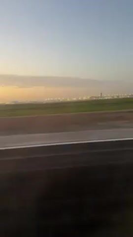 The video shows an airport runway viewed from a moving vehicle, likely an airplane taxiing on the runway. The runway is dark gray asphalt with white lines marking the edge. Adjacent to the runway is a strip of green grass. In the distance, a city skyline is visible under a sky transitioning from a pale blue to a soft orange. The city lights are on, indicating that it is either dawn or dusk. The video is taken at ground level and appears to be handheld or from a vehicle window, as there are minor movements and blurring from the speed.