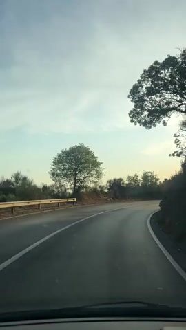 The video shows a view from inside a car driving on a paved road. The road curves to the right, with a white line dividing the lanes. On the left side of the road, there is a guardrail and a grassy slope with a solitary tree. On the right side, there is a rocky incline with some vegetation. The sky is bright with scattered clouds, and the sun is shining, casting long shadows. In the distance, some buildings are visible. The sun's glare is strong, and there are reflections on the car's windshield. The overall impression is that of a scenic drive during either sunrise or sunset.