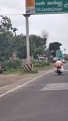 The video captures a roadside scene with a highway and a signpost indicating directions to various towns. A person is riding a motorcycle loaded with luggage, traveling on the highway. In the background, there are trees, buildings, and other vehicles, including a large tanker truck. The sky is overcast, suggesting a cloudy day. The camera appears to be mounted on a moving vehicle, likely following the motorcycle.