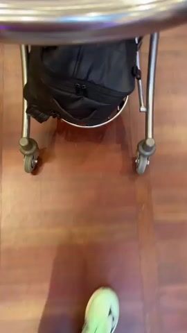 The video shows a first-person perspective of someone walking through an airport with a small luggage cart. The cart holds a black backpack. The floor is made of brown wood planks. The person is wearing light green shoes and light gray pants. Other people are walking in the same direction, some with rolling suitcases. Signs above indicate directions to a transfer center and information. An advertisement for Norwegian Airlines is visible on a large screen.