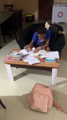 Two young people are engaged in studying or writing indoors in Ijegun, Nigeria.

The first person, wearing a blue t-shirt and black shorts, is seated on a sofa and leaning over a light wooden table. They are actively writing in a notebook with a pen, with an open book positioned beside it. Several other books and notebooks are arranged on the table, along with a box that reads "Smart".

The camera then pans to reveal a second young person lying on their stomach on the tiled floor. This individual, dressed in a white tank top and grey pants, is also focused on writing in a notebook, with books stacked next to them. A pink backpack is visible on the floor in the foreground. The indoor setting features tiled flooring and a white decorative pillar against a light-colored wall. Both individuals appear to be concentrating on their tasks, suggesting a study or homework session.