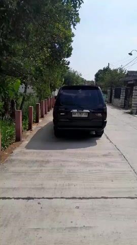 A black SUV drives away from the camera on a paved road. The road is flanked by trees and buildings. The sky is clear and blue.
