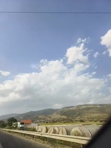 The video is a first-person perspective shot from a vehicle moving along a road. The sky is blue with scattered white clouds. On the right side of the road, there are several large, arched structures covered with a light-colored material, possibly greenhouses or temporary shelters. Beyond these structures, there are hills and mountains in the distance, with some vegetation. On the left side of the road, there are trees, a building, and a power line. The road itself has a white line marking its edge. The camera is angled slightly upwards, capturing the sky and the landscape.