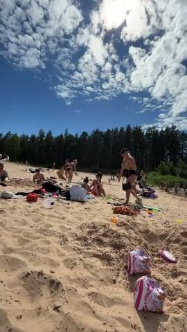 The video shows a sandy beach with a group of people enjoying a sunny day by a body of water. The sky is blue with scattered white clouds. In the foreground, there are footprints in the sand and some bags. Several people are lying on towels or sitting on the sand, some are standing and interacting. Further back, more people are in the water, swimming and playing. A line of trees forms the backdrop to the scene. The water appears calm and inviting.