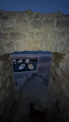 The video shows a view through a stone window in what appears to be an old fort or castle, overlooking a small harbor at dusk. Several small white boats are docked in the harbor. In the distance, across the water, a town is visible, illuminated with lights. There are also several larger boats in the water further away. The sky is a deep blue, indicating either dusk or early evening. The stone of the window appears aged and rough. The perspective is from slightly above, looking down into the harbor. There are at least two people standing on the dock near the boats.