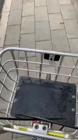 The video shows a first-person view of someone riding a bicycle along a sidewalk. The sidewalk is made of gray rectangular paving stones and runs parallel to a road on the left and a stone wall on the right. The road has a gray asphalt surface and street lights are visible in the background.  A person can be seen in the distance walking on the same sidewalk. The bicycle has a metal wire basket in front. The overall environment appears to be an urban or suburban area with a slight haze or dusk lighting.