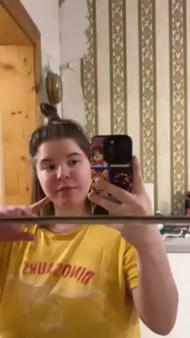 Here's a description of the video:
A young woman is standing in a bathroom, holding her phone in front of her and looking at its screen, which reflects her image in a mirror. She's wearing a yellow t-shirt with a "Dinosaur" print, and her hair is in a messy bun. The bathroom has patterned wallpaper and a wooden door. She makes funny faces, including sticking out her tongue and squinting her eyes.
