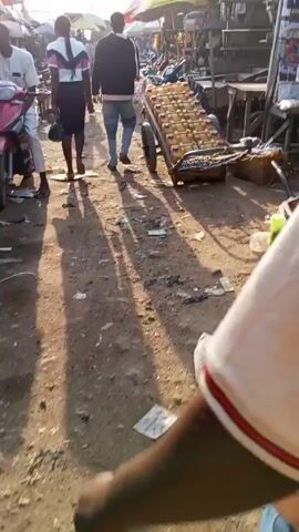 The video shows a person walking through a bustling outdoor market or street. The ground appears to be sandy or dirt-covered, with scattered trash and debris. Other people are walking around, some passing by. The focus is on a cart being wheeled in the same direction as the person filming. The cart is loaded with numerous yellow plastic containers, each with a pink lid. The containers are arranged neatly on the cart. The person filming is wearing a short-sleeved shirt with a white body and a red and white striped trim on the sleeves, along with blue jeans.
