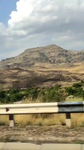 The video shows a moving shot of a mountainous landscape with scattered trees and buildings. A guardrail is visible in the foreground, suggesting the shot is taken from a moving vehicle on a road. The terrain is dry and arid in parts, with patches of green vegetation and some dark areas that could indicate scorched earth. The sky is cloudy.