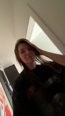 A young woman with long, wet, brown hair wearing a dark t-shirt with a graphic print touches her hair, smiling. The lighting shifts between white, red, and blue. The background includes a white wall, a dark cabinet, and a mirror. Music plays. She continues touching her hair and smiling in the changing lights.