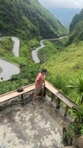 A man in a pink t-shirt, tan shorts, and a gray baseball cap climbs onto the edge of a wooden platform. He is crouched at first, then he sits on the edge with his legs crossed. He smiles at the camera. In the background, a winding mountain road can be seen, with a truck driving along it. The landscape is lush and green.