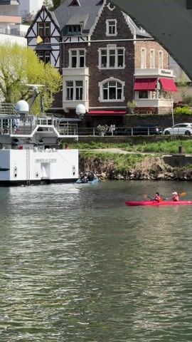 The video shows a river scene with a large white ship named "AMADEUS QUEEN" docked on the left side. Two people are kayaking in a red kayak in the river, paddling towards the right side of the frame. A smaller blue boat with several people in it is positioned between the kayak and the docked ship.

In the background, there's a building with a mix of brown and white walls, featuring a red awning. People can be seen on a waterfront area near the building. A few cars are parked nearby as well.

The camera is positioned under a bridge, as part of the bridge structure is visible at the top of the frame. The river is calm, with gentle ripples on the surface. The sky is clear, suggesting a sunny day.